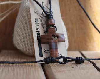 May include: A wooden cross pendant necklace with a heart-shaped cutout. The cross is brown and hangs from a black cord. A tag with the text "Wood & Resin" is attached. The necklace is displayed on a wooden surface.