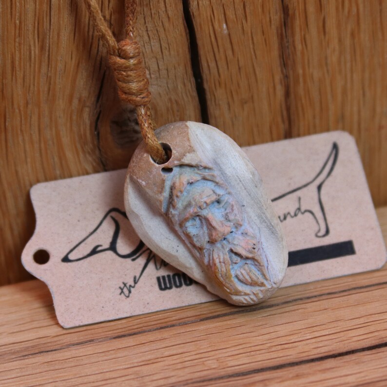 May include: A carved wooden pendant with a face on it, hanging from a brown leather cord. The pendant is light brown and white with a hint of blue. The pendant is hanging over a brown paper tag with the text "the wood hound" on it.