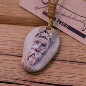 May include: A carved wooden pendant featuring a detailed face with a beard. The pendant is attached to a brown leather cord.