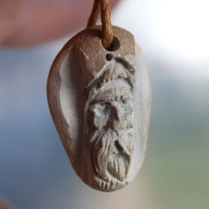 May include: A carved stone pendant with a detailed face of a man with a beard. The pendant is attached to a brown cord.