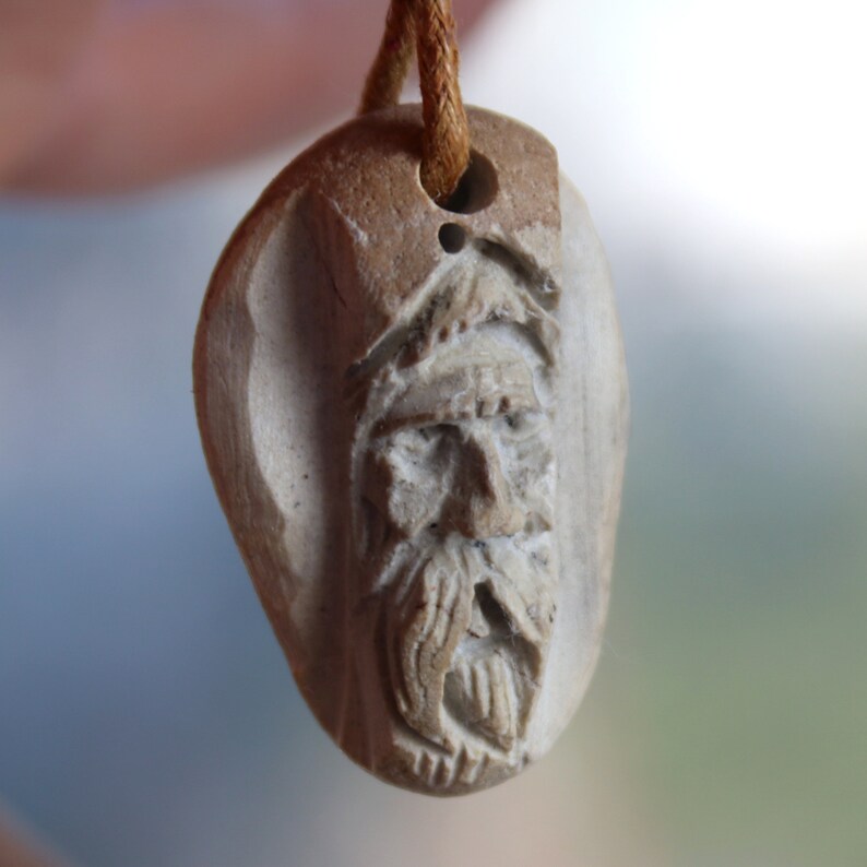 May include: A carved stone pendant with a detailed face of a man with a beard. The pendant is attached to a brown cord.