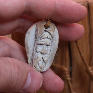 May include: A carved wooden pendant featuring a detailed face with a beard. The pendant is light brown and has a hole at the top for a cord.