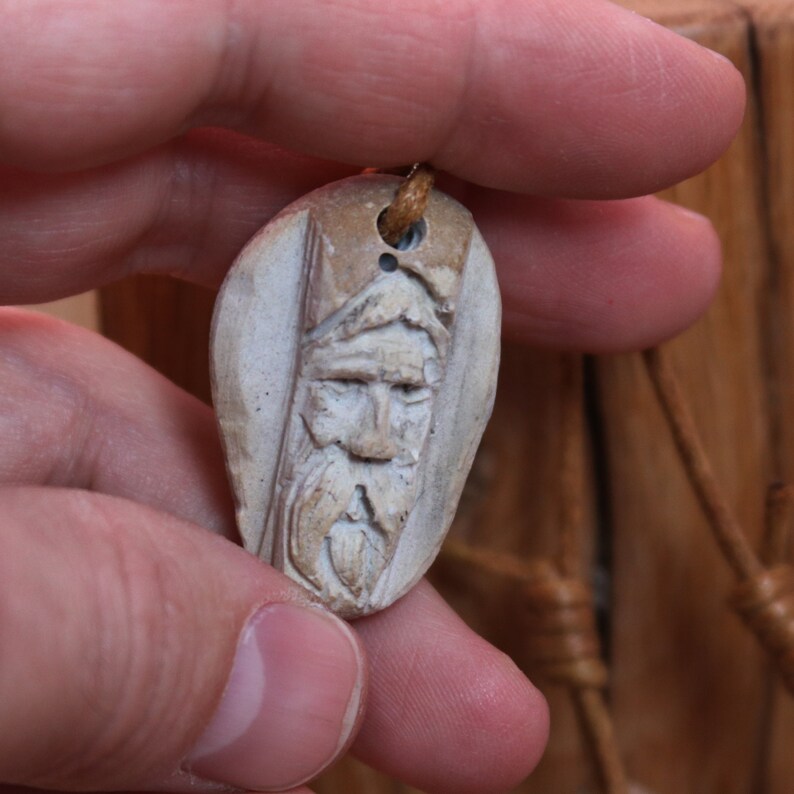 May include: A carved wooden pendant featuring a detailed face with a beard. The pendant is light brown and has a hole at the top for a cord.