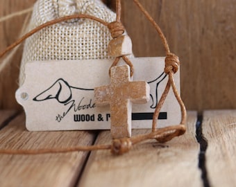 May include: A light brown wooden cross pendant on a brown cord. The cross is knotted to the cord and hangs in front of a small burlap bag and a tag with the text "the Woode WOOD & P". The background is a wooden surface.
