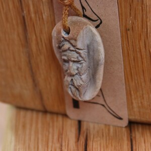 May include: A wooden pendant with a carved face, hanging from a brown cord. The pendant is shaped like a heart and has a detailed carving of a man's face with a beard.