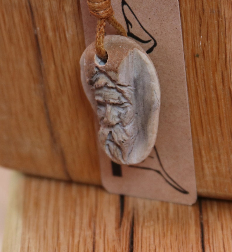 May include: A wooden pendant with a carved face, hanging from a brown cord. The pendant is shaped like a heart and has a detailed carving of a man's face with a beard.