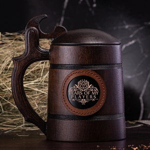 May include: A dark brown wooden beer mug with a handle and a lid. The mug features a leather patch with the text "TEARS OF MY PLAYERS" and a graphic. The handle has a distinctive carved design. The mug is likely for beer or other drinks.