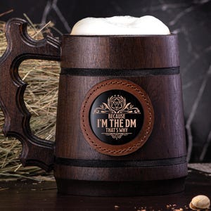 Puede incluir: Una jarra de cerveza de madera marrón con un parche de cuero negro que dice "Because I'm the DM That's Why". El parche tiene una ilustración en blanco y negro de un dado de Dungeons and Dragons.
