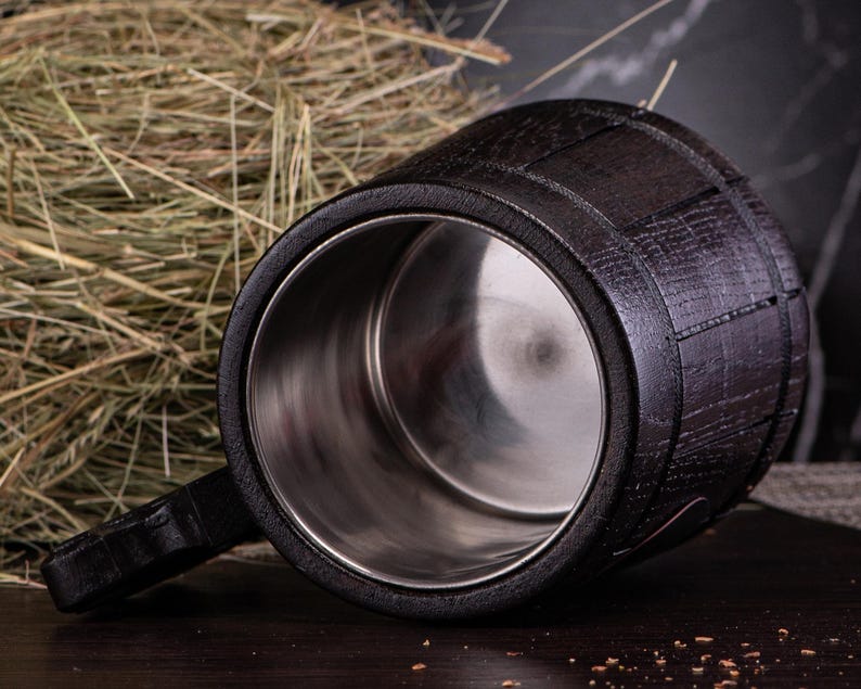 Puede incluir: Un jarro de cerveza de madera marr&oacute;n oscuro con interior de acero inoxidable. El jarro tiene un asa y un dise&ntilde;o similar a un barril. El jarro est&aacute; sobre una superficie de madera oscura.