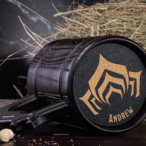 Puede incluir: Un jarro de cerveza de madera oscura con un dise&ntilde;o decorativo y el nombre "ANDREW" en dorado. El jarro tiene forma de barril con un asa y est&aacute; sobre un fondo de heno.