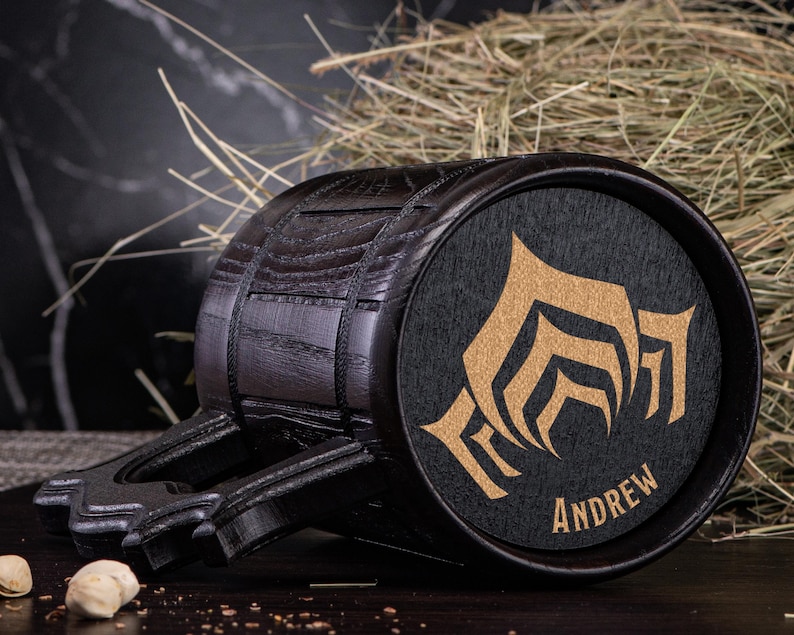 Puede incluir: Un jarro de cerveza de madera oscura con un dise&ntilde;o decorativo y el nombre "ANDREW" en dorado. El jarro tiene forma de barril con un asa y est&aacute; sobre un fondo de heno.