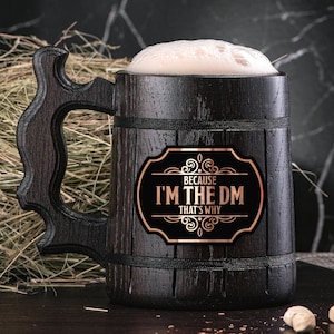 May include: A dark brown wooden beer stein with a handle. The stein has a black and gold sticker that says "Because I'm the DM That's Why".