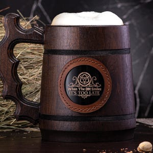 May include: A brown wooden beer stein with a black leather patch that says "When The DM Smiles It's Too Late". The stein has a foam head on top of the beer.