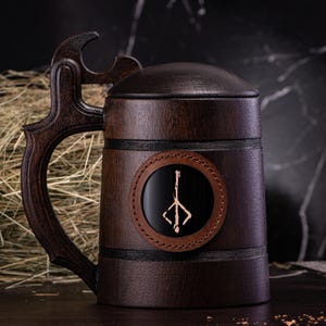 May include: A dark brown wooden beer stein with a lid and a large handle. The stein features a circular emblem with a stylized design on the front. The handle has a unique, decorative shape. The stein is designed to hold beverages.