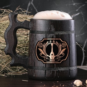 May include: A dark brown wooden beer stein with a black and gold sticker featuring a lute and the word "BARD". The stein is filled with a light brown liquid.