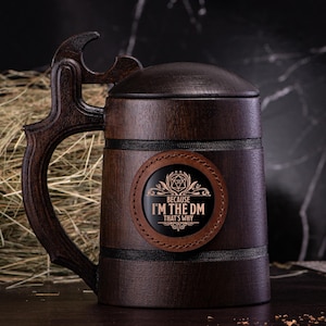 Puede incluir: Un jarro de cerveza de madera marrón oscuro con asa curva y tapa. El jarro tiene un parche de cuero con el texto "BECAUSE I'M THE DM THAT'S WHY" y un gráfico de fantasía. Ideal para los aficionados a los juegos de rol.