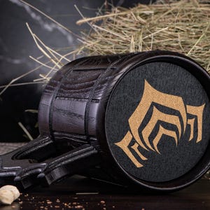 Puede incluir: Un jarro de cerveza de madera oscura con un asa negra y un emblema circular con un dise&ntilde;o dorado. El jarro tiene forma de barril con bandas met&aacute;licas y una base plana. El emblema est&aacute; sobre un fondo negro.