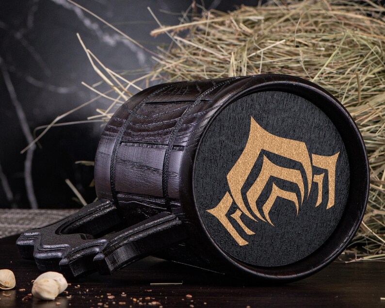 Puede incluir: Un jarro de cerveza de madera oscura con un asa negra y un emblema circular con un dise&ntilde;o dorado. El jarro tiene forma de barril con bandas met&aacute;licas y una base plana. El emblema est&aacute; sobre un fondo negro.