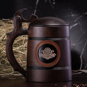 Puede incluir: Una jarra de cerveza de madera marrón con un parche de cuero que dice "Dungeon Master" y un símbolo de dado dorado.