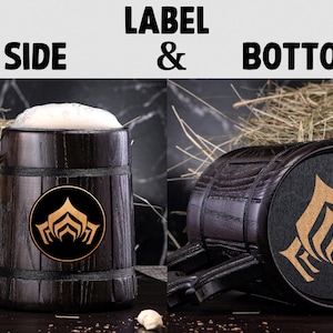 Puede incluir: Taza de cerveza de madera oscura, llena de cerveza y espuma. La taza tiene un asa decorativa y un emblema circular. La parte inferior de la taza tambi&eacute;n es visible, con un emblema a juego.