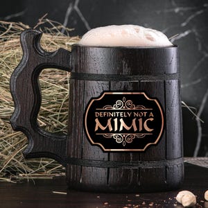 May include: A dark brown wooden beer stein with a black and gold sticker that says "Definitely Not A Mimic". The stein is filled with a light brown liquid and has a foam head.