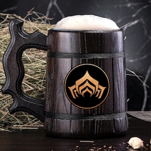 Puede incluir: Una jarra de cerveza de madera oscura con un asa grande, llena de cerveza y espuma. La jarra tiene un emblema dorado y negro en la parte delantera. El fondo incluye heno y una superficie oscura.