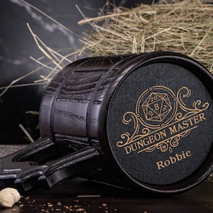 May include: A black wooden mug with a gold design that says "Dungeon Master" and "Robbie". The mug has a 20-sided die graphic and a decorative scroll design.