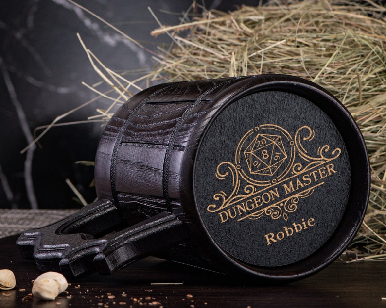 May include: A black wooden mug with a gold design that says "Dungeon Master" and "Robbie". The mug has a 20-sided die graphic and a decorative scroll design.