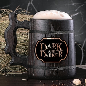 Könnte beinhalten: Ein dunkelbraunes, hölzernes Bierkrug mit einem schwarzen und goldenen Etikett, auf dem "DARK AND DARKER" steht. Der Krug ist mit einer hellbraunen Flüssigkeit gefüllt und hat einen weißen Schaumkopf.