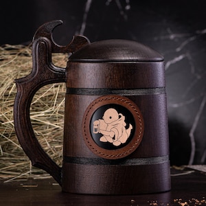 May include: A dark brown wooden beer stein with a matching lid and handle. The stein features a circular emblem with a light brown illustration of a figure holding a mug. The handle has a decorative cutout.
