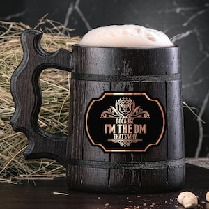 Könnte beinhalten: Ein dunkelbraunes, hölzernes Bierkrug mit einem schwarzen und goldenen Aufkleber, auf dem "Because I'm the DM That's Why" steht. Der Aufkleber zeigt einen stilisierten 20-seitigen Würfel.