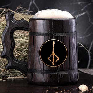 May include: A dark wooden beer stein filled with beer and foam. The stein has a large handle and a gold emblem on the front. The emblem is a stylized symbol on a black background. The stein is sitting on a dark surface.