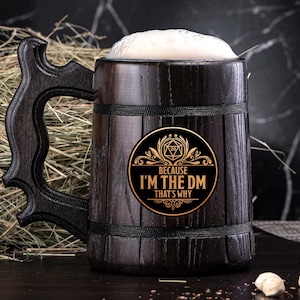 Könnte beinhalten: Ein dunkelbraunes, hölzernes Bierkrug mit einem schwarzen Henkel und einem goldenen Aufkleber mit der Aufschrift "Because I'm the DM That's Why". Der Aufkleber zeigt einen 20-seitigen Würfel und ein dekoratives Design.
