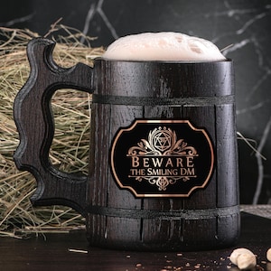Könnte beinhalten: Ein dunkelbraunes, hölzernes Bierkrug mit einem schwarzen und goldenen Aufkleber, auf dem "Beware the Smiling DM" steht. Der Krug hat einen Henkel und ist mit einer hellbraunen Flüssigkeit gefüllt.