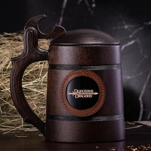 May include: A dark brown wooden beer mug with a lid and a handle featuring an integrated bottle opener. The front of the mug displays a circular leather patch with the Dungeons & Dragons logo printed in white text and a sword graphic.