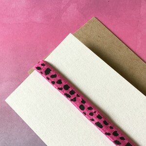 May include: A pink elastic band with black spots rests on a white card and a brown card, set against a pink gradient background. The band is a vibrant pink with irregular black spots.