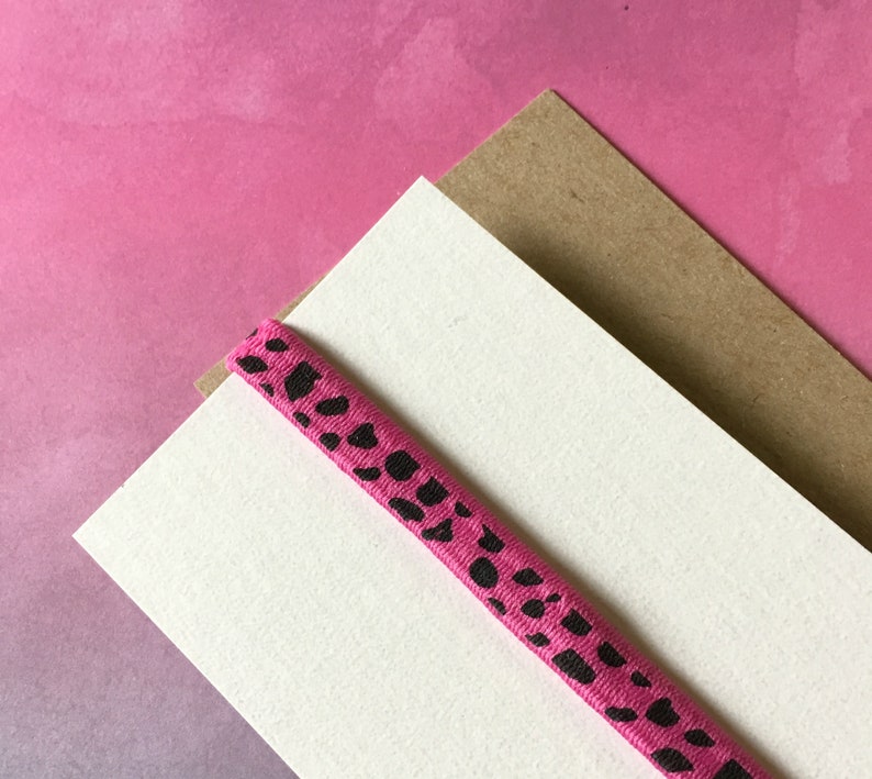 May include: A pink elastic band with black spots rests on a white card and a brown card, set against a pink gradient background. The band is a vibrant pink with irregular black spots.