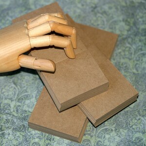 May include: A stack of square, brown paper cards. A wooden articulated hand is positioned above the cards, with its fingers slightly curved. The cards are stacked in a neat pile, with several other stacks visible.