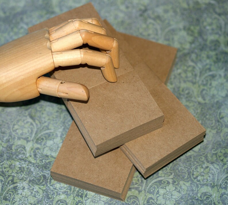 May include: A stack of square, brown paper cards. A wooden articulated hand is positioned above the cards, with its fingers slightly curved. The cards are stacked in a neat pile, with several other stacks visible.