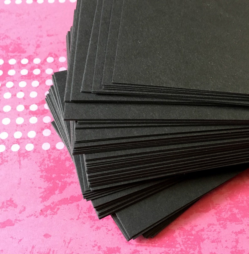 Pu&ograve; includere: Una pila di carte quadrate nere. Le carte sono disposte ordinatamente, con bordi visibili. Lo sfondo &egrave; una superficie rosa strutturata con pois bianchi. Le carte sembrano essere fatte di carta spessa e opaca.