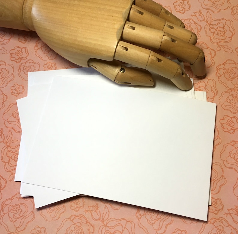 Puede incluir: Una pila de tarjetas blancas en blanco con esquinas redondeadas, que se encuentran sobre una superficie de estampado floral rosa. Una mano de madera est&aacute; sosteniendo la tarjeta superior.
