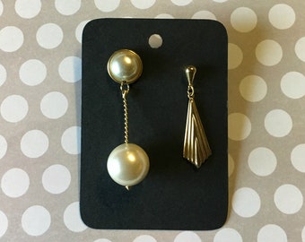 May include: A pair of gold earrings with a pearl drop and a geometric design. The earrings are displayed on a black card.