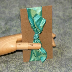May include: A teal satin ribbon tied in a bow around a brown rectangular tag. The ribbon has a slight sheen and is knotted in the center. The tag is held by a wooden hand.