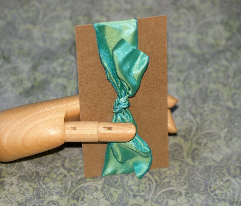 May include: A teal satin ribbon tied in a bow around a brown rectangular tag. The ribbon has a slight sheen and is knotted in the center. The tag is held by a wooden hand.