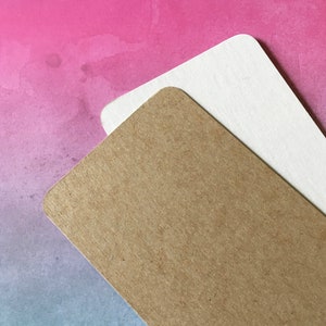 May include: Two rectangular paper tags with rounded corners. One tag is a light brown color, and the other is white. The tags are layered on top of each other, with the brown tag in the foreground. The background is a watercolor wash of pink, purple, and blue.