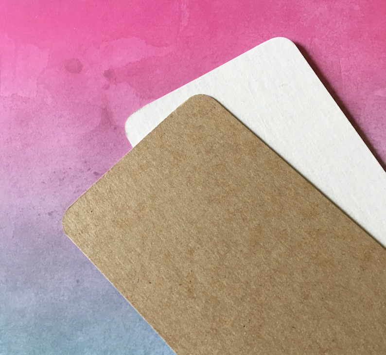 May include: Two rectangular paper tags with rounded corners. One tag is a light brown color, and the other is white. The tags are layered on top of each other, with the brown tag in the foreground. The background is a watercolor wash of pink, purple, and blue.