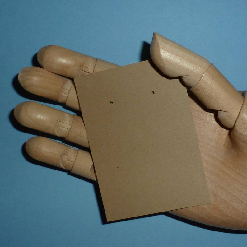 May include: A brown cardboard earring display card with two holes for earrings. The card is held in a wooden hand.