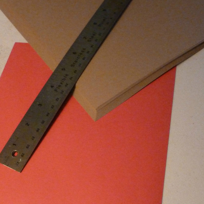 May include: A brown piece of paper with a red piece of paper underneath. A metal ruler with the words "Flexible Stainless Steel Ruler" printed on it is placed on top of the brown paper.