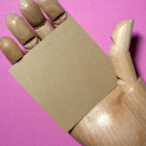 May include: A brown paper card held by a wooden hand model against a pink background.