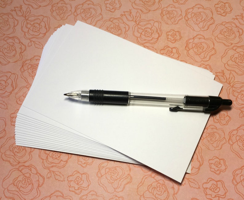 Puede incluir: Una pila de tarjetas blancas en blanco con un bol&iacute;grafo negro en la parte superior. Las tarjetas est&aacute;n sobre un fondo rosa con un patr&oacute;n floral.